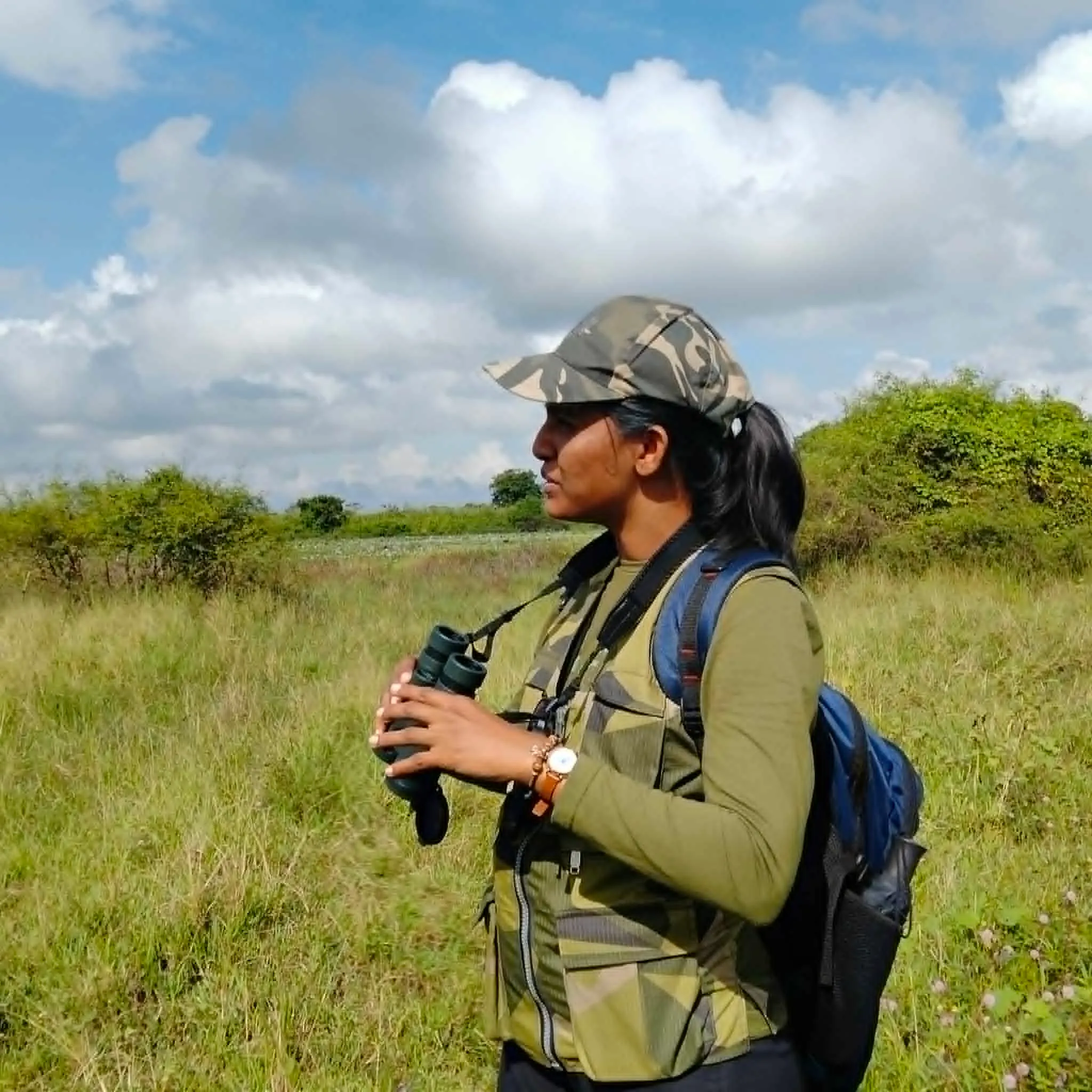 Standing in open grassland with binoculars raised, wearing a camouflage cap and vest, scanning a leopard-range landscape