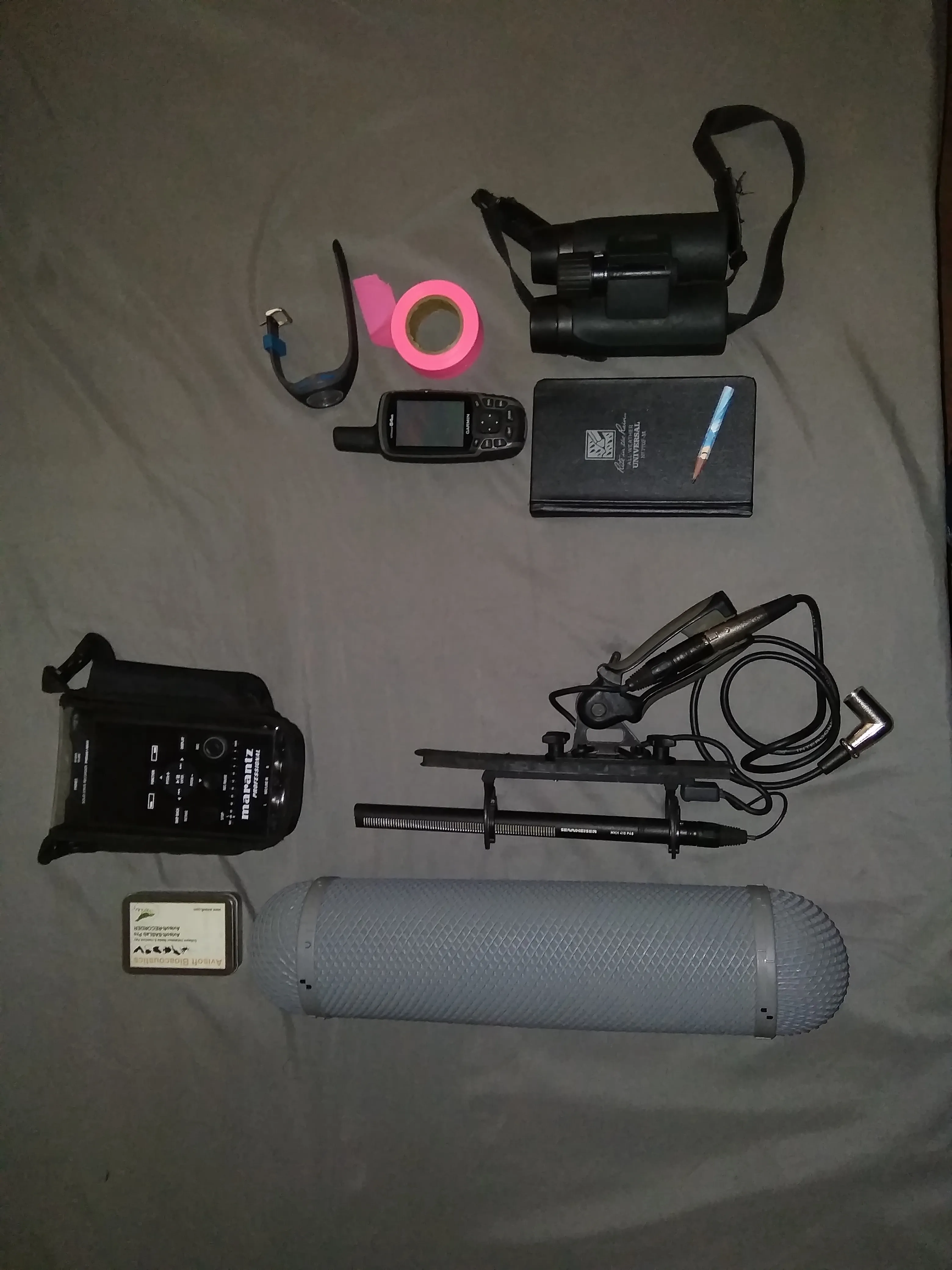 Field equipment laid out on a groundsheet: binoculars, a GPS unit, a field notebook, a Marantz audio recorder, and a Sennheiser shotgun microphone with windshield