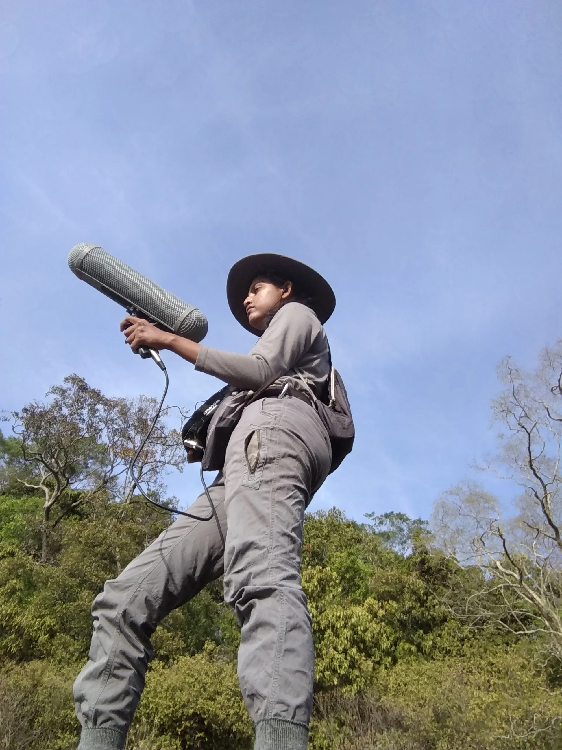 Low-angle photograph of a field researcher aiming a shotgun microphone skyward, wearing a wide-brimmed hat against a bright sky