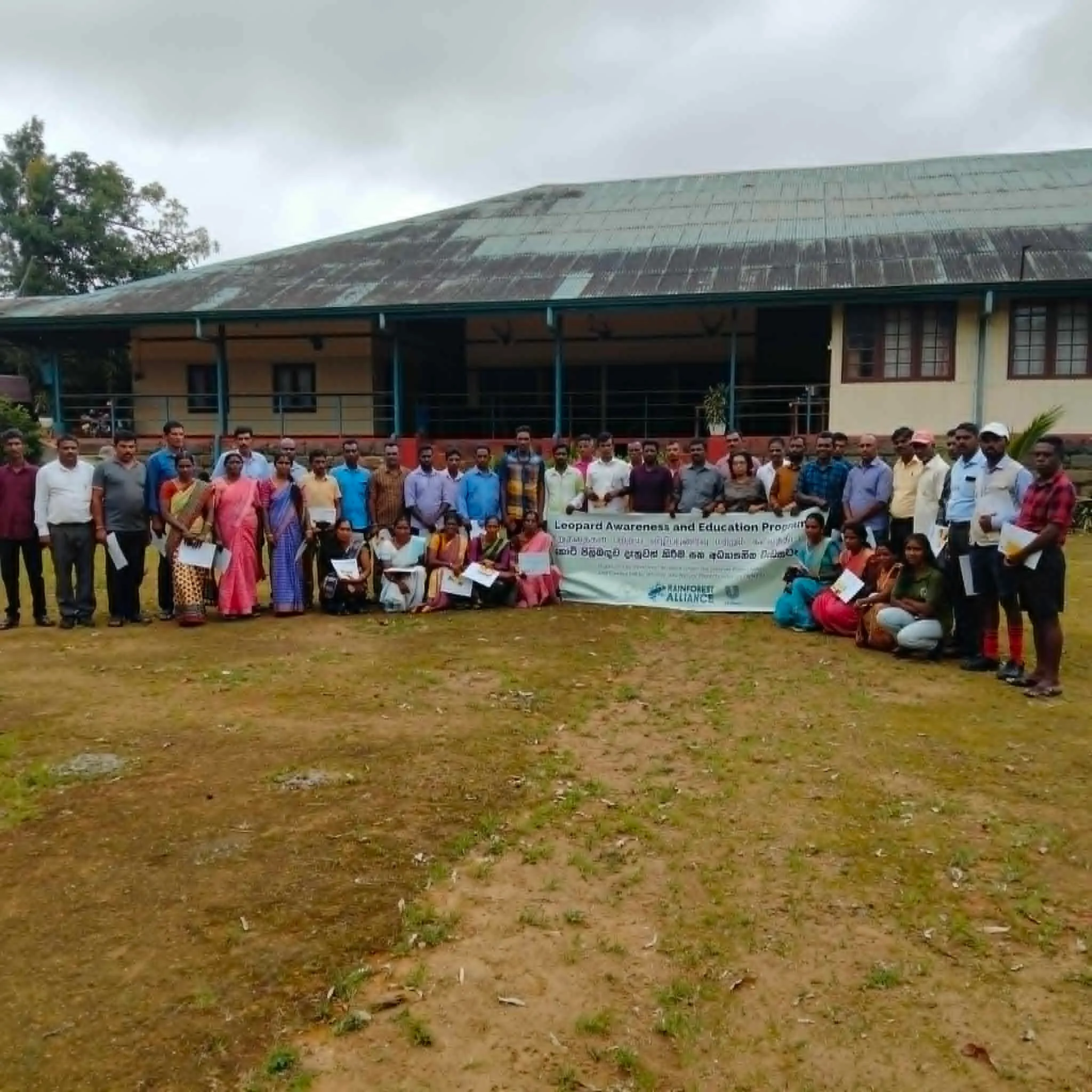 A large group of tea estate workers and community members standing outdoors in front of an estate building, holding leaflets, behind a banner reading "Leopard Awareness and Education Programme"