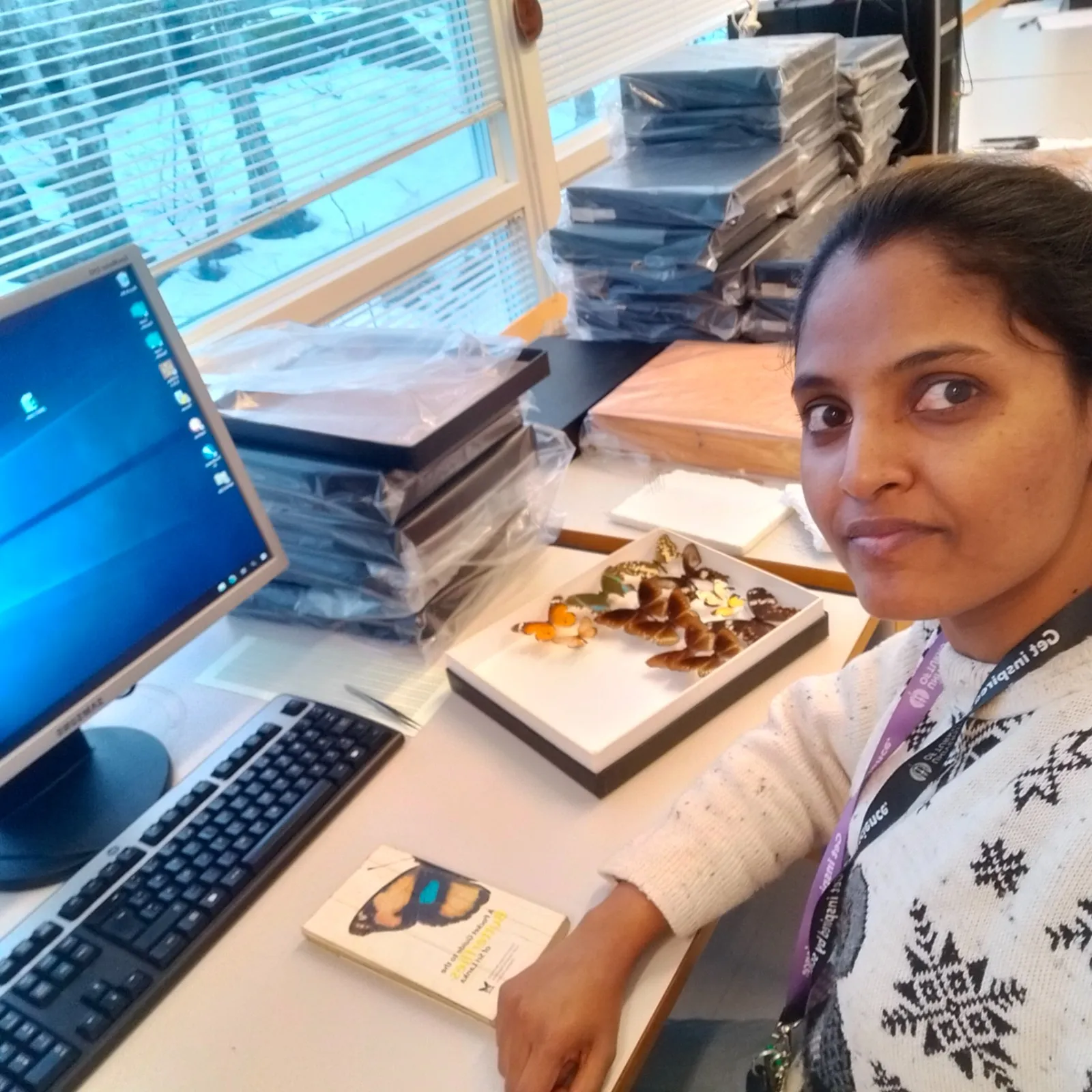 Seated at a desk in the Turku natural history collection room, beside a computer and a tray of pinned butterflies, with stacks of specimen boxes behind and snow visible through the window