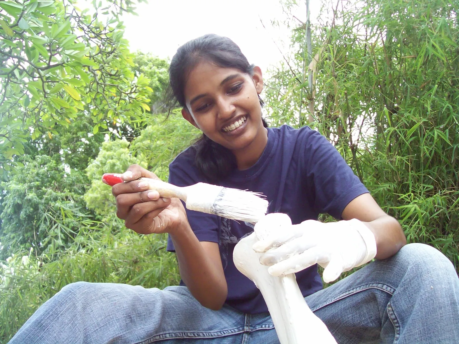 Sitting outdoors in gloves, using a brush to clean a large animal leg bone, with green foliage behind